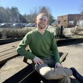 A man sits outside on a picnic table wearing a green shirt and beige pants, smiling at the camera.