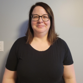 A woman with long, brown hair stands against a grey wall wearing a black shirt and glasses.