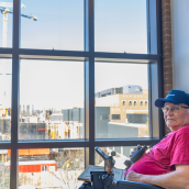 A man sits in a wheelchair with a window behind him with a view of a construction site and a crane.