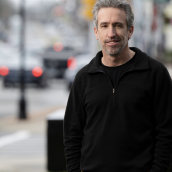 A man wears a black jacket and shirt with brown pants and is standing outside with vehicles on the road in the background, smiling at the camera.