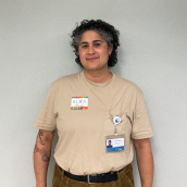 A person with short, curly hair, wearing a beige shirt and brown pants stands in front of a white wall, smiling at the camera.