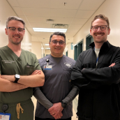 Three men stand together smiling in the hallway of a healthcare facility.