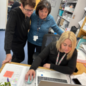 3 healthcare workers, 2 standing and 1 sitting in front of a laptop in an office.