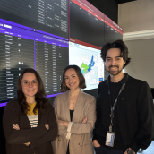 Pictured are 2 women and 1 man (R). Smiling with 4 large television screens behind them.