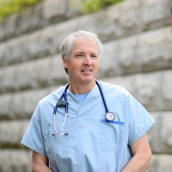 Picture of a male physician in light blue scrubs with stethoscope around his neck. He has grey short hair and is smiling. The picture is taken outside with a rock wall scenery outdoors. 