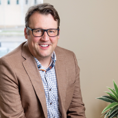 Image of a man with light brown hair and glasses. He is wearing a brown sports coat and blue and white checkered shirt. He is smiling largely and he is seated in an office.