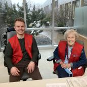 A man and woman sit side-by-side in chairs at a healthcare facility wearing red vests and smiling at the camera.