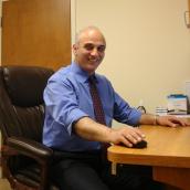 A man sits in a chair wearing a blue-collared shirt and a red tie in front of a computer screen and is smiling at the camera.