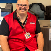 A bald man in a red vest, black short-sleeve shirt sits in a healthcare facility smiling at the camera.