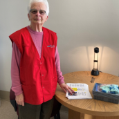 A woman with short, white hair wears a red vest and pink long-sleeve shirt stands in a hospital room beside a table, smiling at the camera.