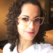 A woman with long, brown, curly hair wears glasses and a white shirt, smiling at the camera.