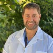 A man stands outside with a white lab coat and is smiling at the camera.