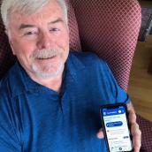An older man with gray hair and a trimmed beard sits in a red patterned armchair, smiling at the camera while holding a smartphone that displays a healthcare app with options for finding care and services.
