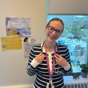 Image of a woman in a blue and white striped button sweater. She's wearing glasses, hair tied neatly back and smiling. She has a stethoscope and lanyard around her neck. The photo is taken in a office.