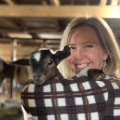 Photo of a woman with blonde woman wearing a brown and white checkered jacket holding a baby goat in her arms.