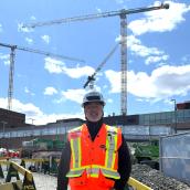 A man stands in front of a construction site wearing an orange safety vest and hard hat.