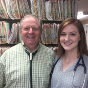 Photo of a man (L) and woman (R) standing in a room with medical charts/folders behind them. They both are smiling standing next to each other very professional with stethoscope's around their necks.