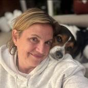 A woman with her dog in a home. She has blonde hair and is wearing a white hoodie. Her dog is black, brown and white and their head is on her shoulder. 