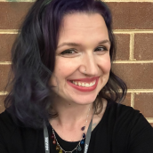 A woman with black short curly hair and a purple streak at the front. She is wearing a black t-shirt, a colourful necklace and a black lanyard.
