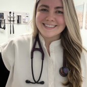 Photograph of woman wearing white scrub top and a stethoscope around her neck, smiling with long blonde hair. Behind her, the wall of a medical office can be seen with a few medical supplies hanging up. 