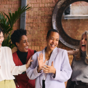 Photo of 6 women smiling and looking at each other. Mix of black and white women in a room with brown brick behind them, a plant and a round dark framed mirror.