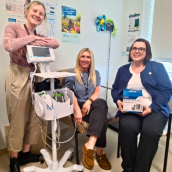 Picture of 3 women. Woman on the left is leaning over an automatic blood pressure machine, woman in the middle is getting her blood pressure taken and there is a woman in scrubs on the right side of the picture holding a box. They are all smiling largely. 