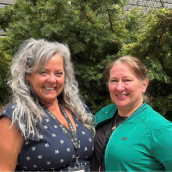 Two women stand side-by-side in front of a green leafy bush, smiling at the camera.