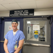 Picture of a man in blue scrubs and scrub hat standing in front of operating room doors. There is a sign above his head that reads "Restricted area operating rooms authorized personnel only."