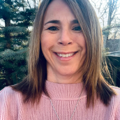 Photo of a woman with dark brown shoulder length hair smiling largely. Has a pink sweater and a necklace with a large pendant around her neck. The photo is taken outside with trees in the background.