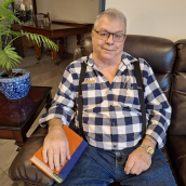Man with glasses and short hair sits in a chair wearing a plaid shirt, suspenders and jeans.