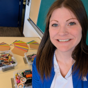 A smiling woman fills the right side of the frame, taking a cheerful selfie in what looks like a classroom or creative workspace. Behind her, a tabletop is neatly arranged with craft materials. 