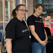 Two women indoors, standing side by side, both wearing black t-shirts with the text “Bay Gals” printed on them.  