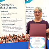 In an indoor setting, a smiling woman holds a certificate standing in front of banners related to community health boards. 