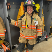 Image of a female firefighter in full gear standing next to a open closet with firefighter gear hanging up.