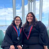photo of two women in graduation regalia sitting in front of a window