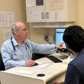 A man wearing a stethoscope and blue shirt sits at a desk across from another person and is pointing to a computer screen.