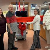 Four people pose with an ultrasound machine with a ribbon tied around it to celebrate its arrival at a local hospital.