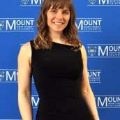 A smiling woman is standing in front of a blue backdrop covered with the Mount Saint Vincent University logo and text. 