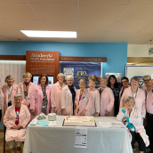 Image of a large group of women and one male in the picture. All people are wearing light pink lab style long jackets. There are 2 women sitting and the rest are standing - all around a table with a white table cloth and a cake sitting on top.
