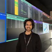 Man standing in front of digital screens wearing a black shirt, lanyard, and has dark hair and a beard.