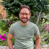 Close up image of a man standing outside in front of a garden. He has his hands in his pockets, wearing glasses and smiling showing his teeth in a short sleeve light green shirt.