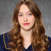 A formal portrait of a young woman in an indoor setting wearing a graduation gown looking directly at the camera. 