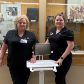 Two healthcare workers in black scrubs are standing inside a building with glass display cabinets behind them. They are standing with a portable computer between them.