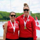 Two women on a podium wearing sunglasses, red shirts, and both have a medal around their neck. 