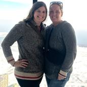 Two women are standing close together and smiling warmly for the camera. The background opens out into a bright cityscape and water view.