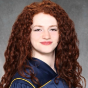 A studio-style portrait of a young woman with long curly red hair wearing a graduation gown. 