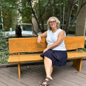 Debbie Burris (woman wearing a white top and navy skirt, short light hair and glasses. Sandals on her feet) sitting on a bench in the Sheila Gloade Memorial Healing Garden.