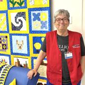 A smiling woman stands indoors beside a brightly colored quilt display, resting one hand on the back of a blue couch. 