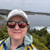 A woman stands outdoors wearing a ball cap, sunglasses and a raincoat, smiling at the camera.