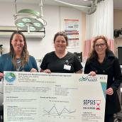 Three health care staff stand in an emergency department, smiling and holding a large poster about the Northern Zone Sepsis Care Directive initiative. Medical equipment, an overhead exam light and a trauma management checklist are visible in the background.